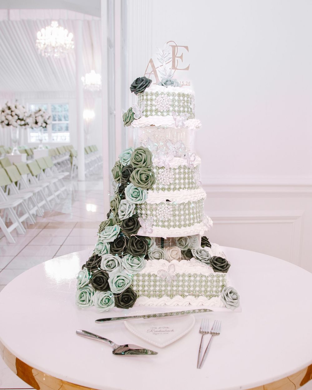 Elegant six-tier wedding cake with green and white roses, pearl details, and monogram topper on a white table.