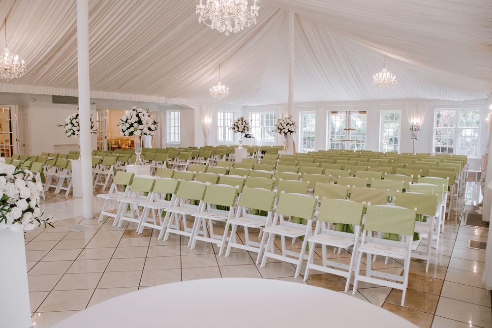 Elegant wedding ceremony venue with white folding chairs, green cushions, chandelier lighting, and floral arrangements under a draped tent.
