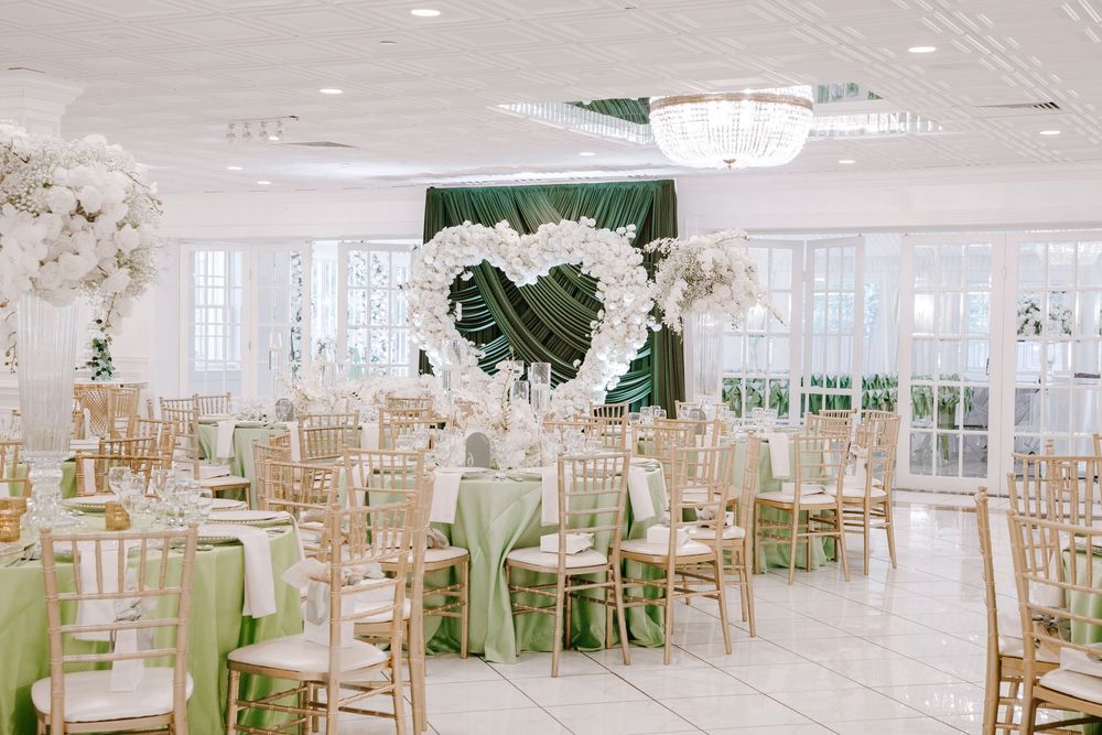 Elegant wedding reception hall with gold chiavari chairs, sage green linens, and a floral heart arch centerpiece.