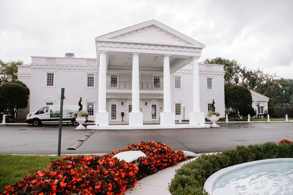 White colonial mansion with grand pillars, red flowers, and fountain on cloudy day.