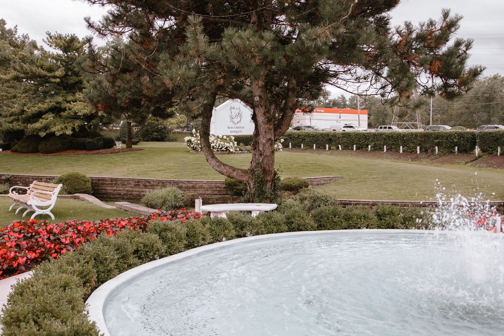 Serene garden fountain with red flowers, white bench, lush trees, and manicured lawn in a peaceful park setting.
