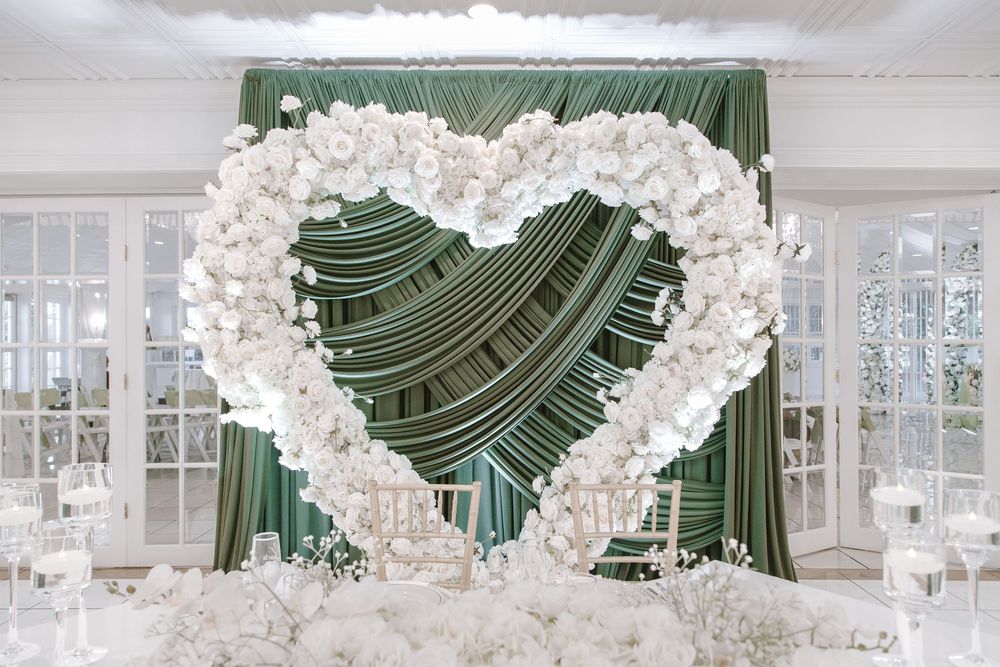 Elegant wedding arch decorated with white flowers in a heart shape against green draped backdrop.