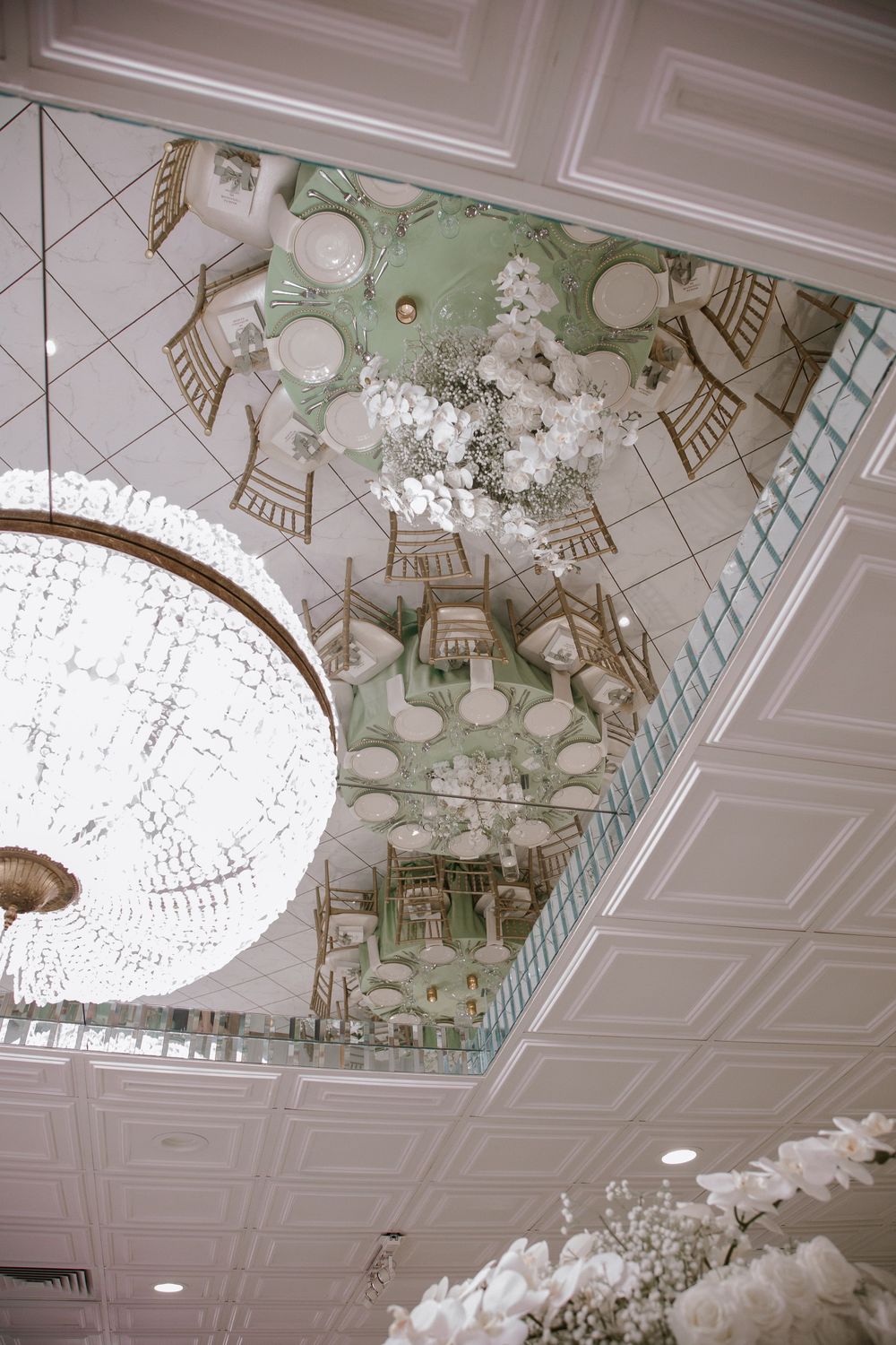 Elegant ballroom ceiling with crystal chandelier, mirrored panels, and green mural reflecting ornate decor and white floral arrangements.