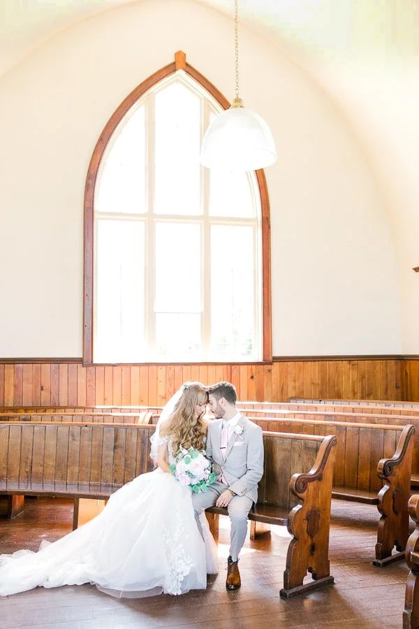 Bride and groom share an intimate moment seated on a church pew, bathed in natural light from an arched chapel window.