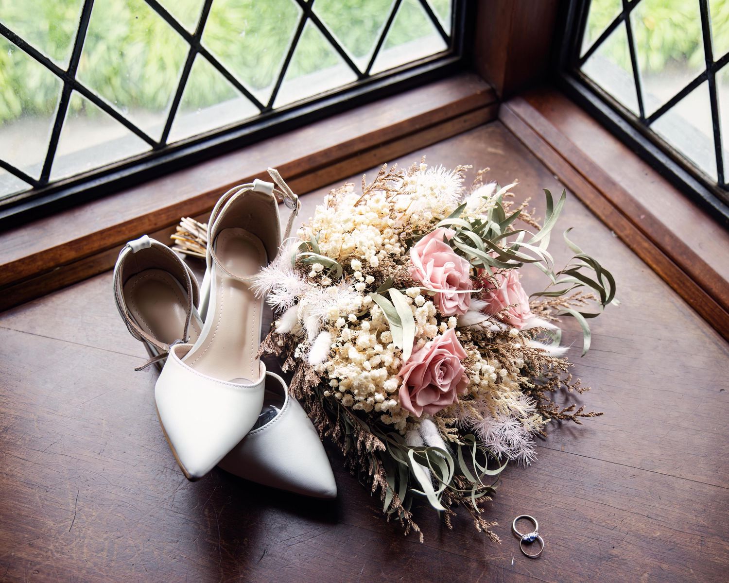 White bridal heels and wedding rings beside a romantic bouquet of pink roses and dried flowers on a wooden windowsill.