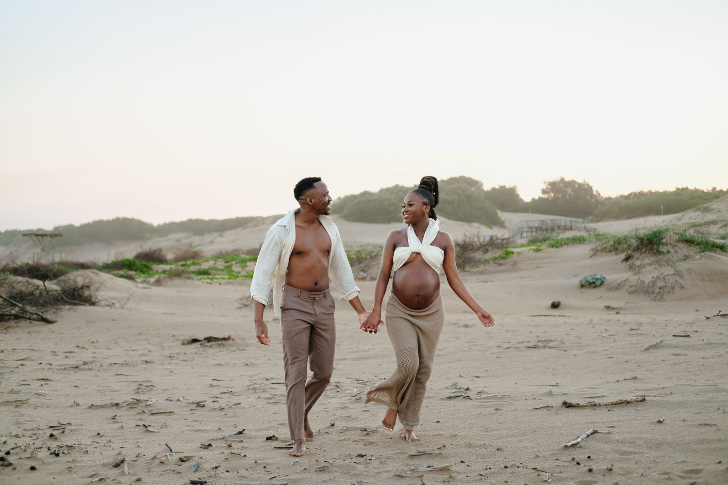 Maternity couple holding hands while walking along the beach at sunset