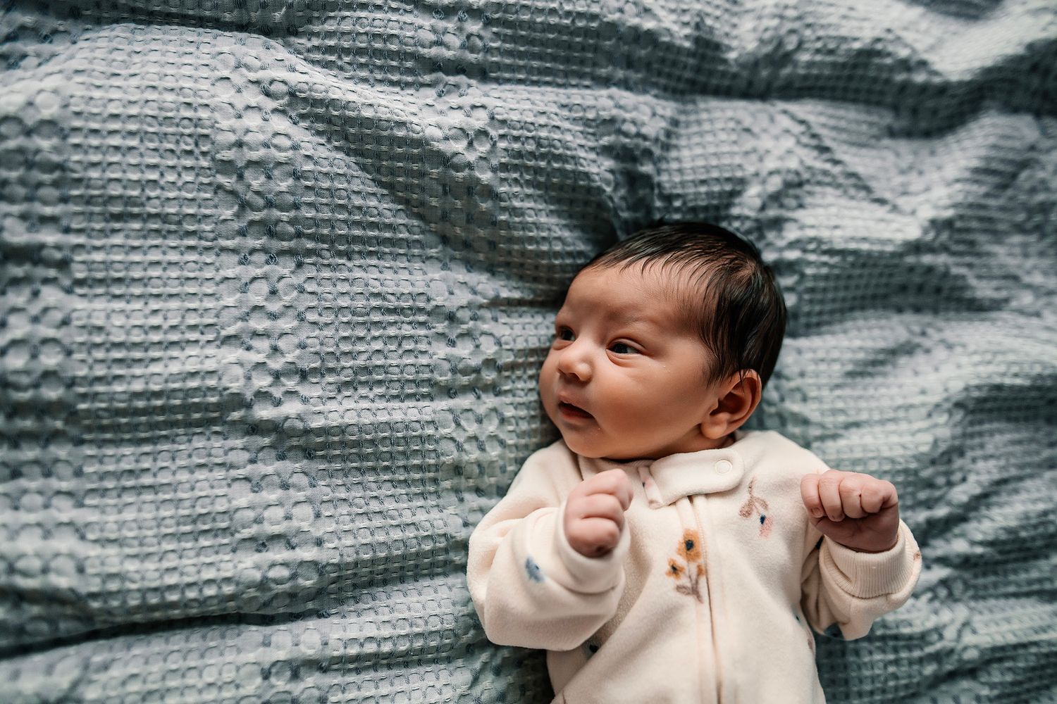 A newborn baby in a white onesie lying on a blue textured blanket, looking up with tiny fists raised.