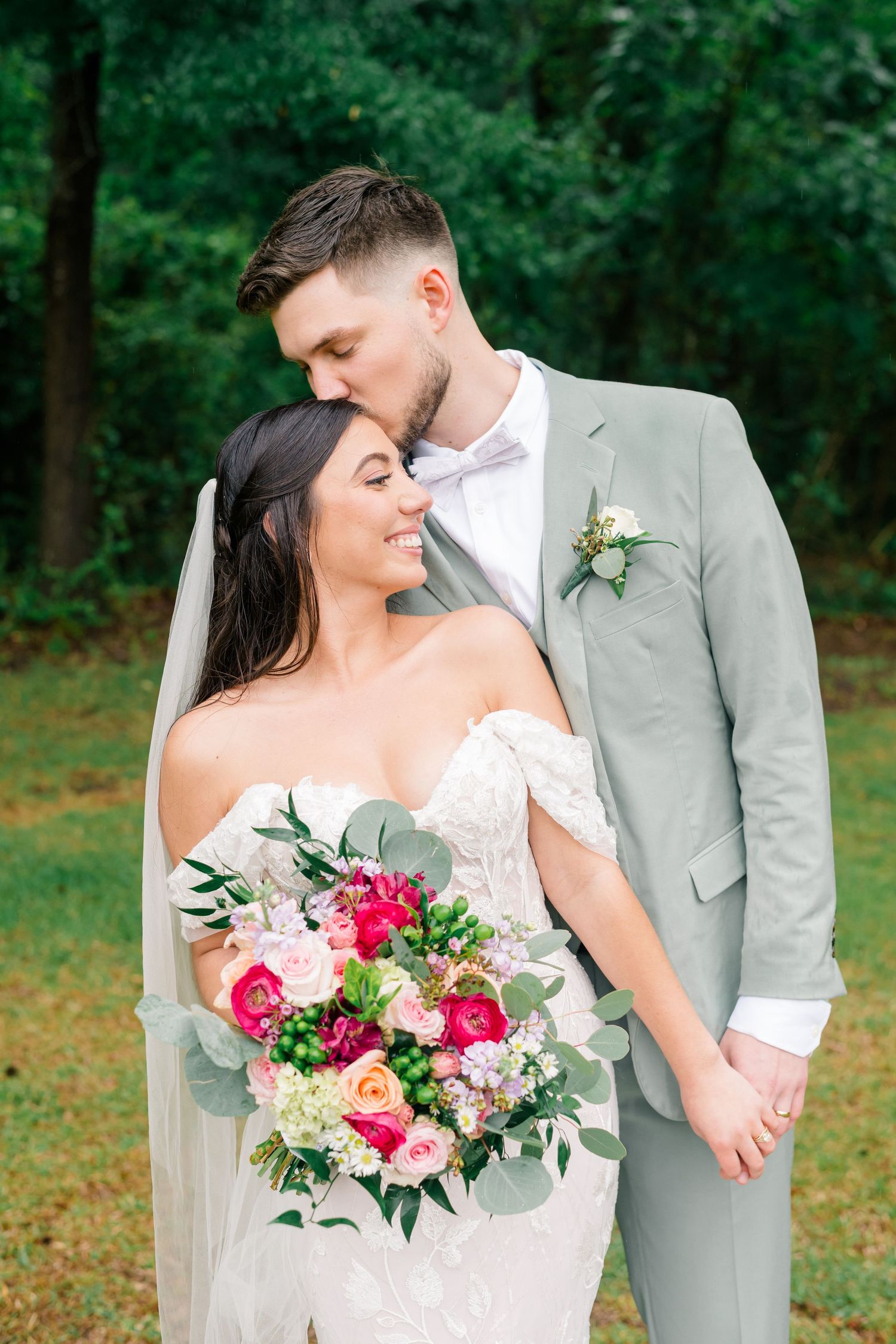 Groom in sage suit kisses bride wearing lace off-shoulder gown holding colorful bouquet outdoors.