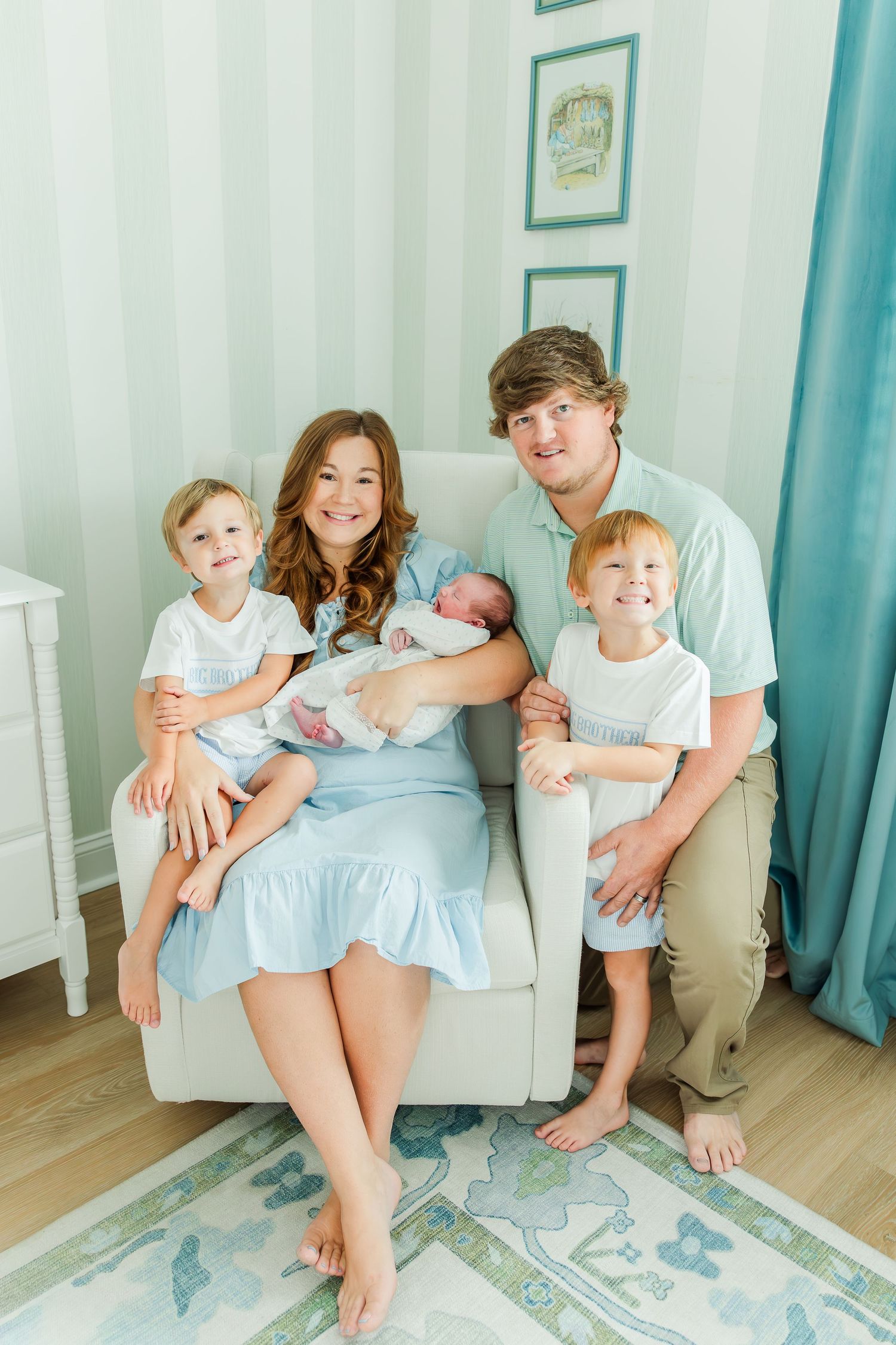 Happy family of five posing in a soft blue nursery, parents with two young boys and a newborn baby.