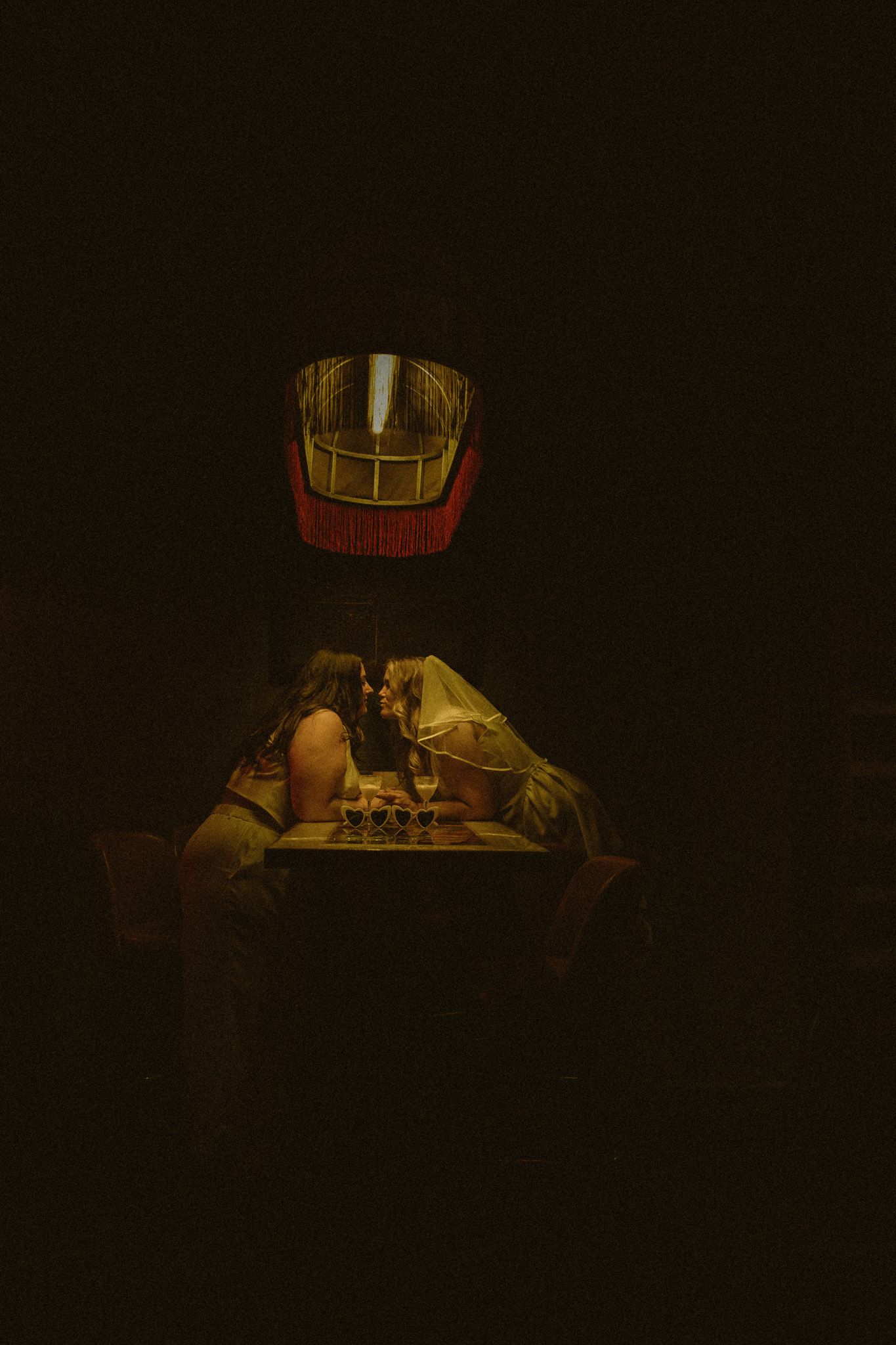 Two brides sharing a romantic kiss under a decorative fringed lamp in a dimly lit venue.