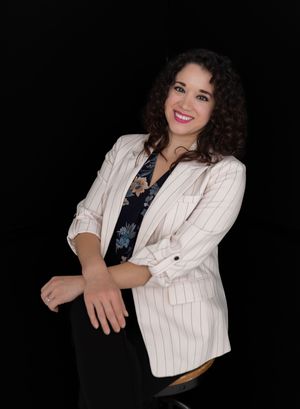 Smiling woman with curly hair wearing a white pinstripe blazer over floral top, posing confidently against black background.