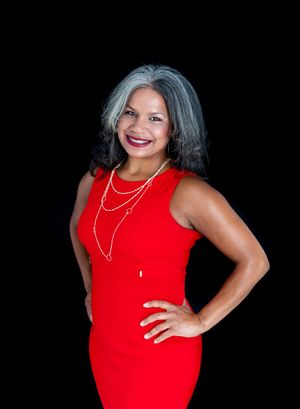 Woman in red sleeveless dress with gray hair and pearl necklace posing confidently against black background.