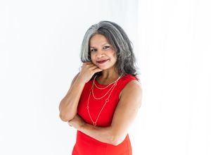 A confident woman in a red sleeveless dress with gray hair and gold necklaces poses against a white background.