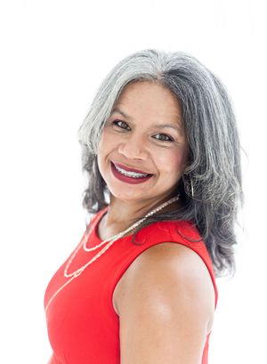 Smiling middle-aged woman with gray streaked hair wearing a red sleeveless top and pearl necklace against white background.