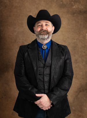Man in black cowboy hat, leather jacket, blue shirt, and star bolo tie poses confidently for a portrait photo.