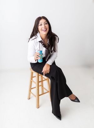 Woman in white shirt and black pants sits on a stool, smiling and holding a beverage can.