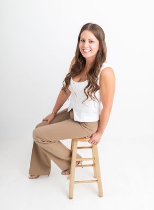 A smiling brunette woman sits on a wooden stool wearing a white top and tan wide-leg pants against a white background.