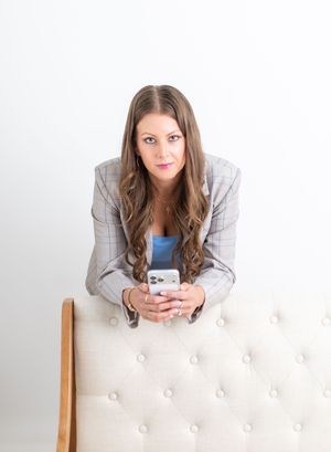 Professional woman in gray blazer leaning on tufted chair holding smartphone, looking at camera.