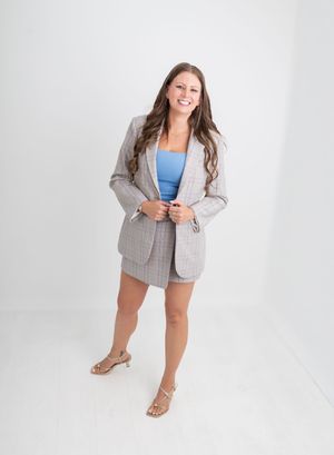Woman smiling in plaid blazer and skirt set with blue top and strappy heels on white background.