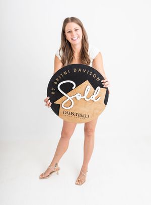 Smiling woman holding a round black and gold Sold sign for Britni Davison at Dawsch & Co Real Estate.