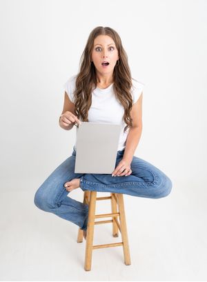 Surprised woman sitting on wooden stool holding laptop, wearing white tee and jeans, against white background.