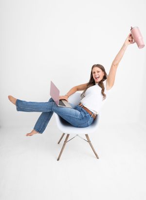 Woman in jeans celebrating with laptop and pink mug raised in air while sitting on modern white chair.