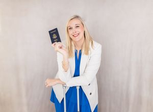 Smiling blonde woman in white blazer and blue dress holding a US passport, posing with luggage.