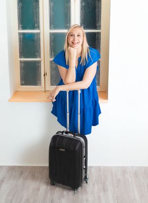 Smiling blonde woman in blue dress leaning on black carry-on luggage near a window.