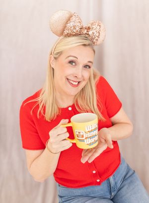 Smiling blonde woman in red shirt holding Walt Disney World mug wearing rose gold Minnie Mouse ears headband.