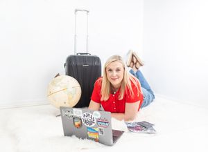 Blonde woman in red shirt lying on white fur rug with laptop, globe, and suitcase, travel blogger setup.