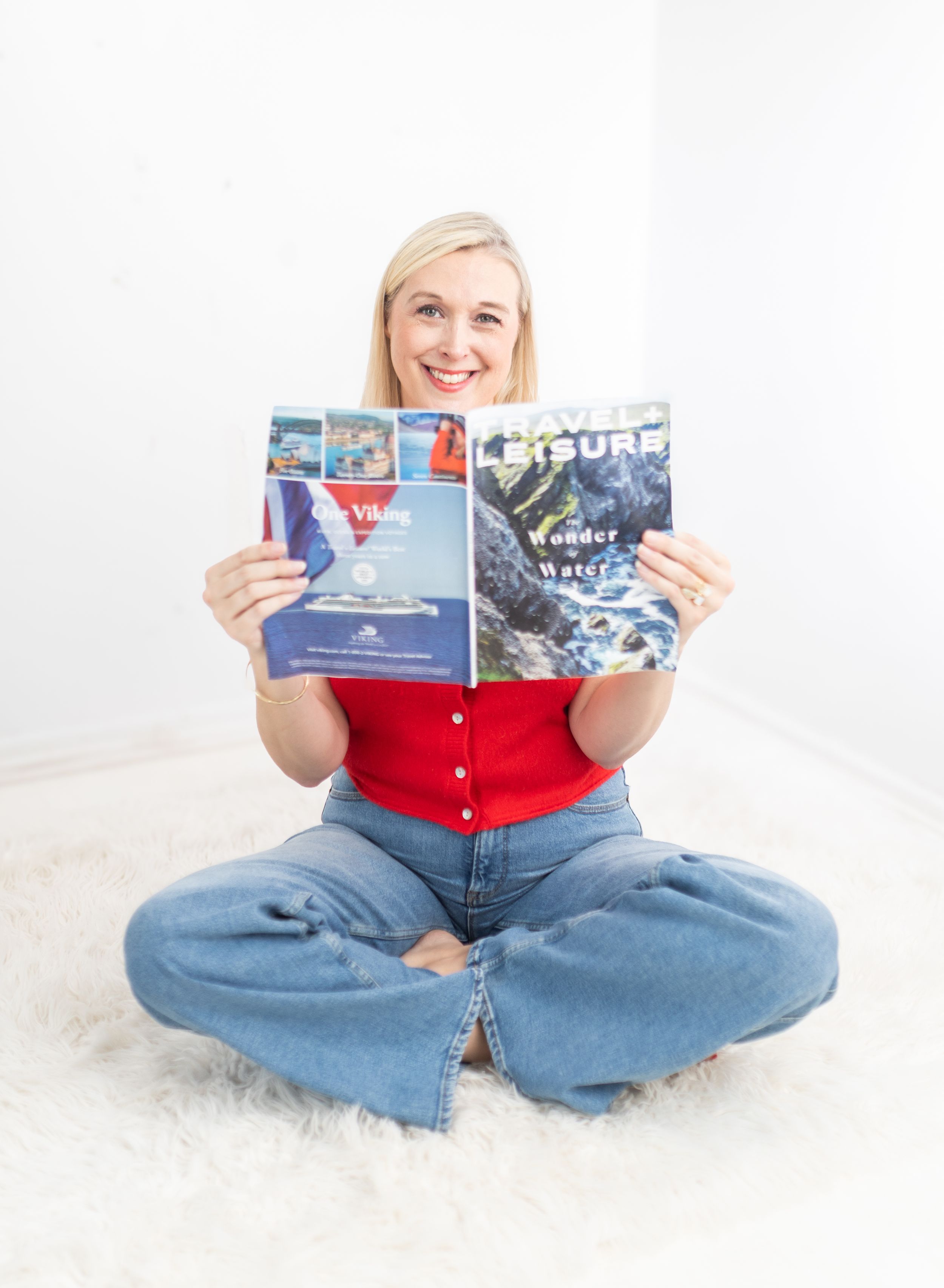 Smiling blonde woman sitting cross-legged on white rug, holding open a Travel Leisure magazine, wearing red top and jeans.