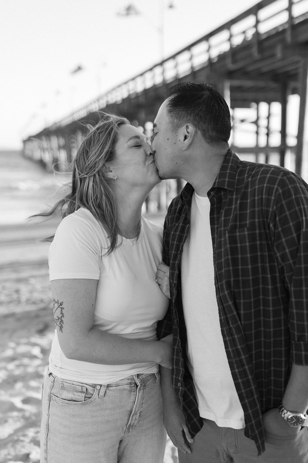 A classic California pier is in the background of a couple kissing for their engagement session on a California beach.