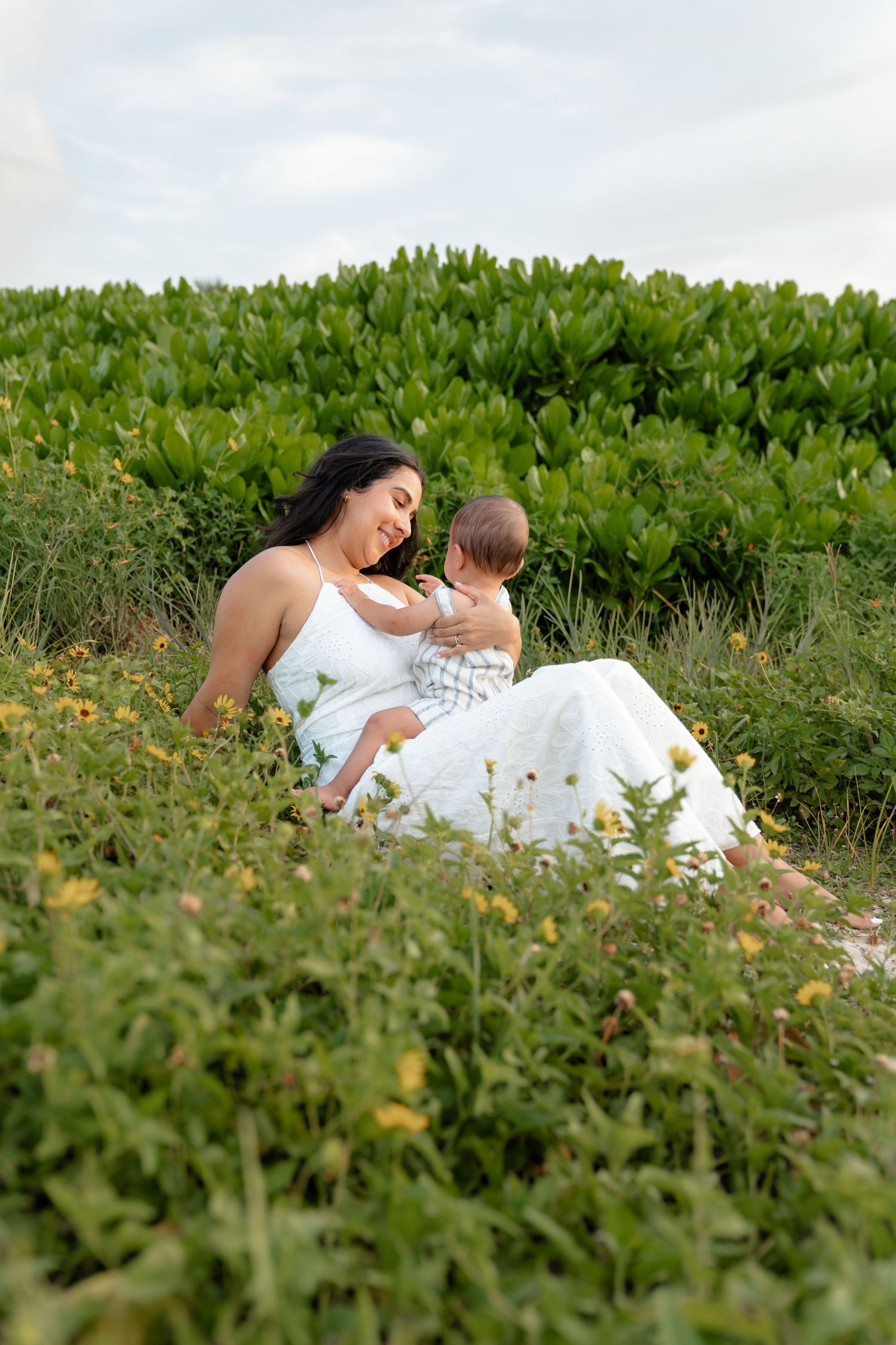 A mother in a white dress sits in a lush green field, lovingly cradling her baby outdoors.
