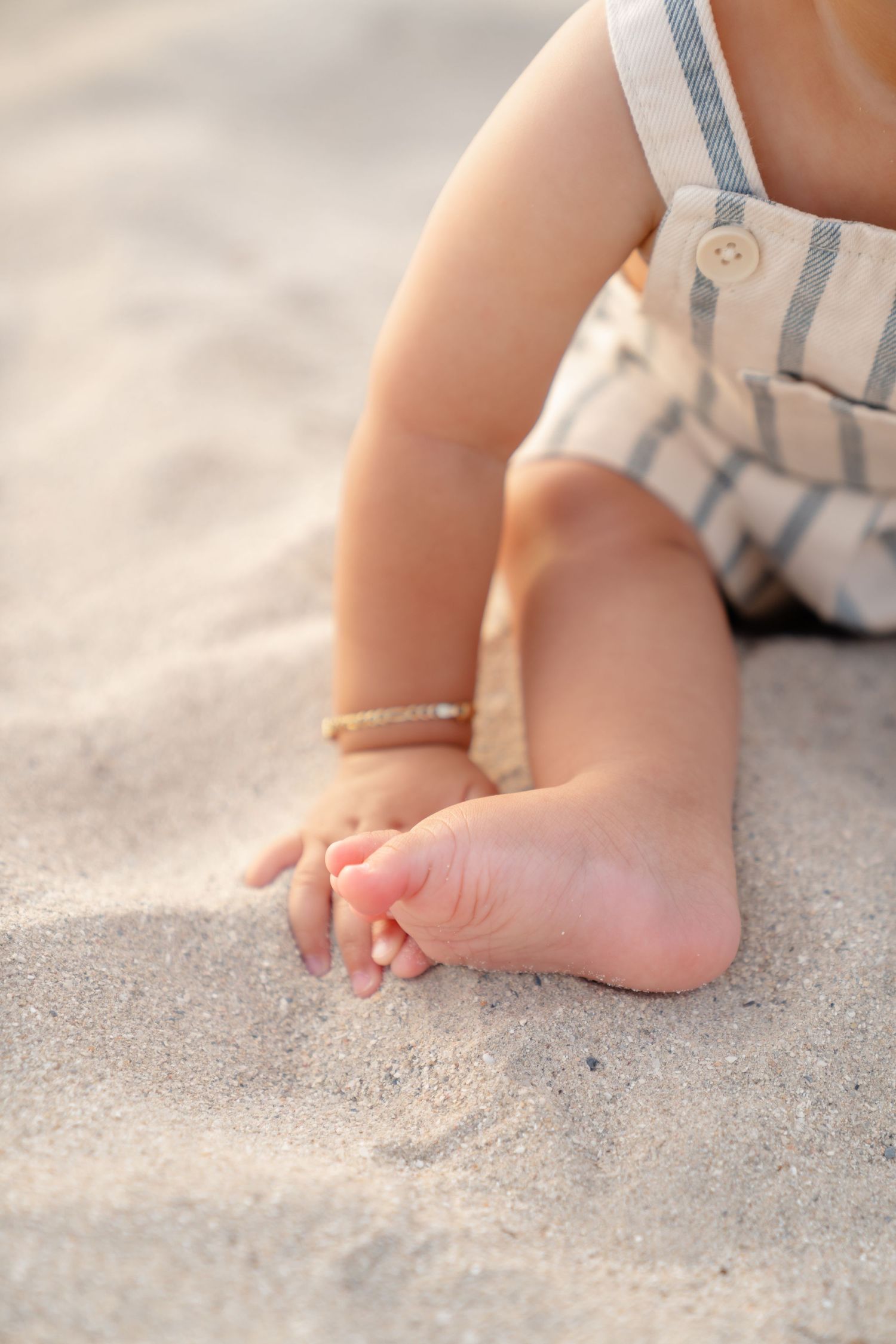 Close-up of baby feet in sand at the beach, wearing striped overalls and a delicate gold anklet bracelet.