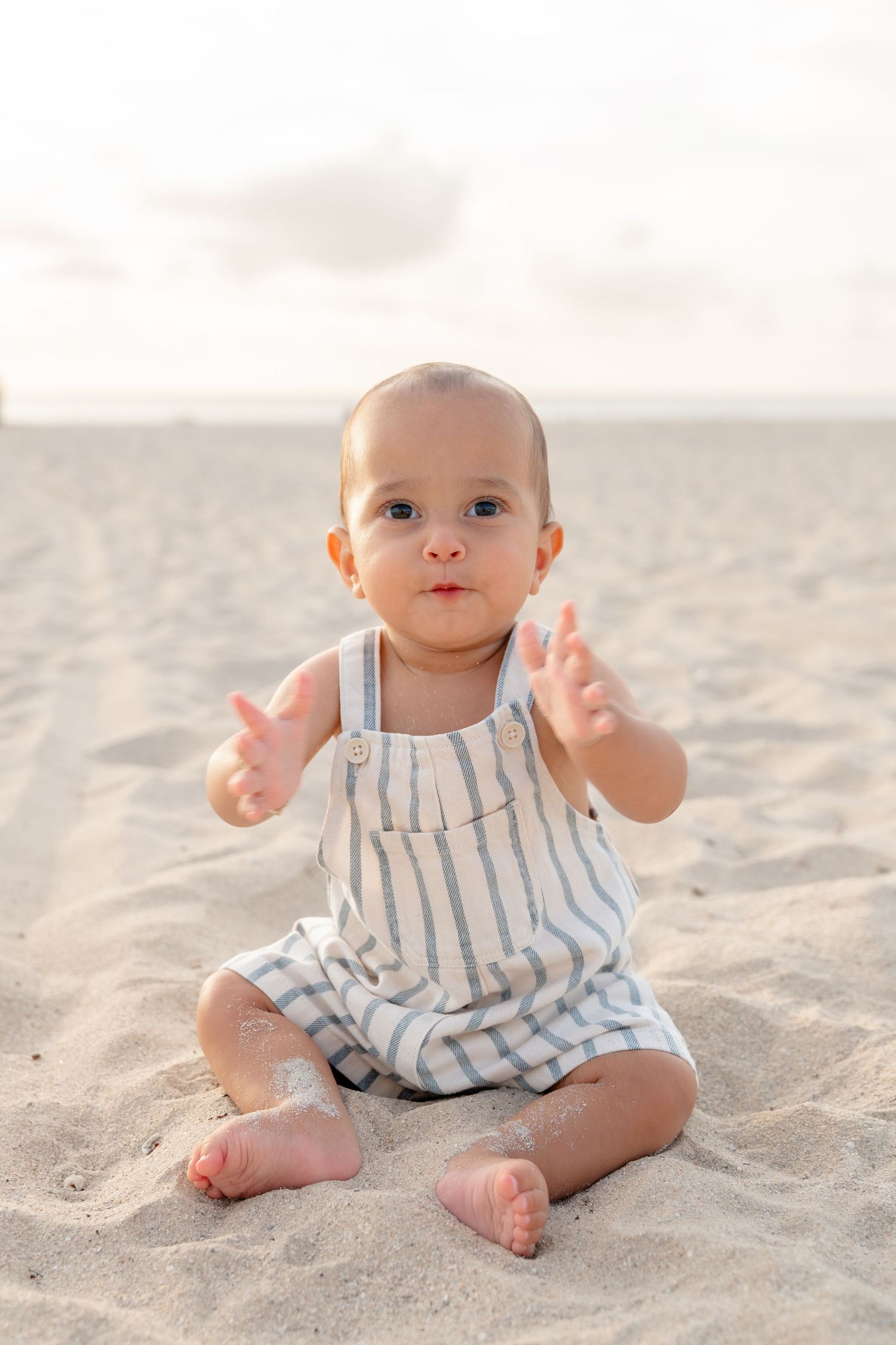 Cute baby in striped overalls sitting on sandy beach, reaching out hands toward camera on a sunny day.