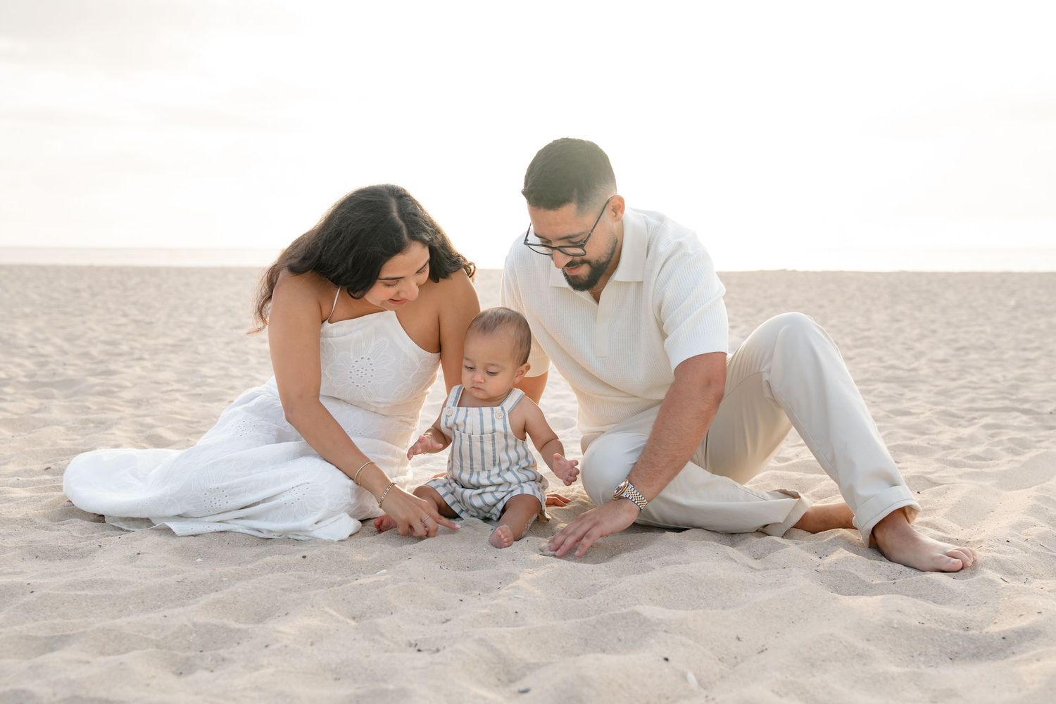 Family of three sitting on sandy beach, parents in white outfits with baby in striped romper, soft natural light.