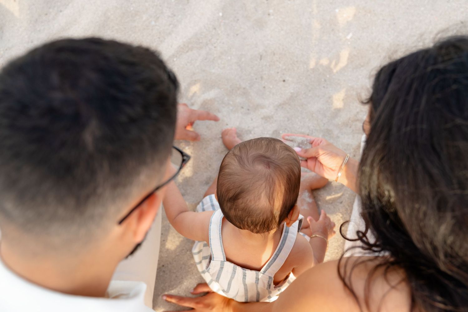 Parents sitting on sandy beach with their baby, viewed from above in warm golden light.