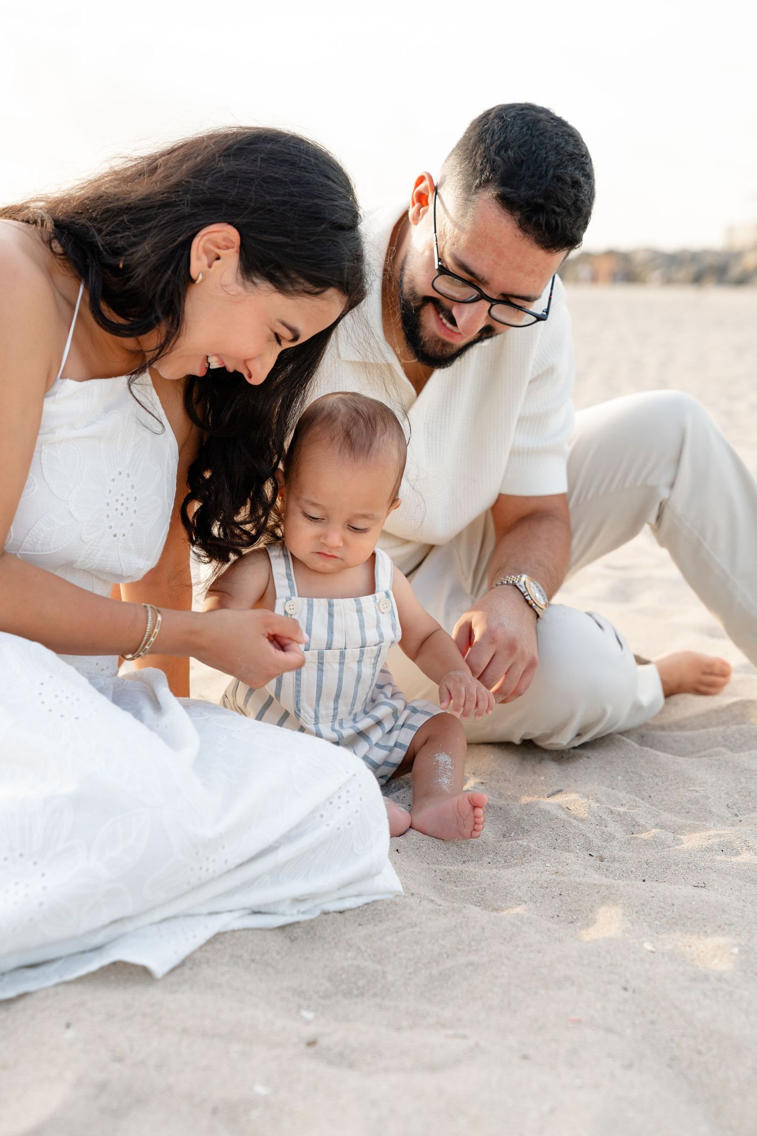 A young family with a baby sits on sandy beach, parents smiling as their infant explores the sand.
