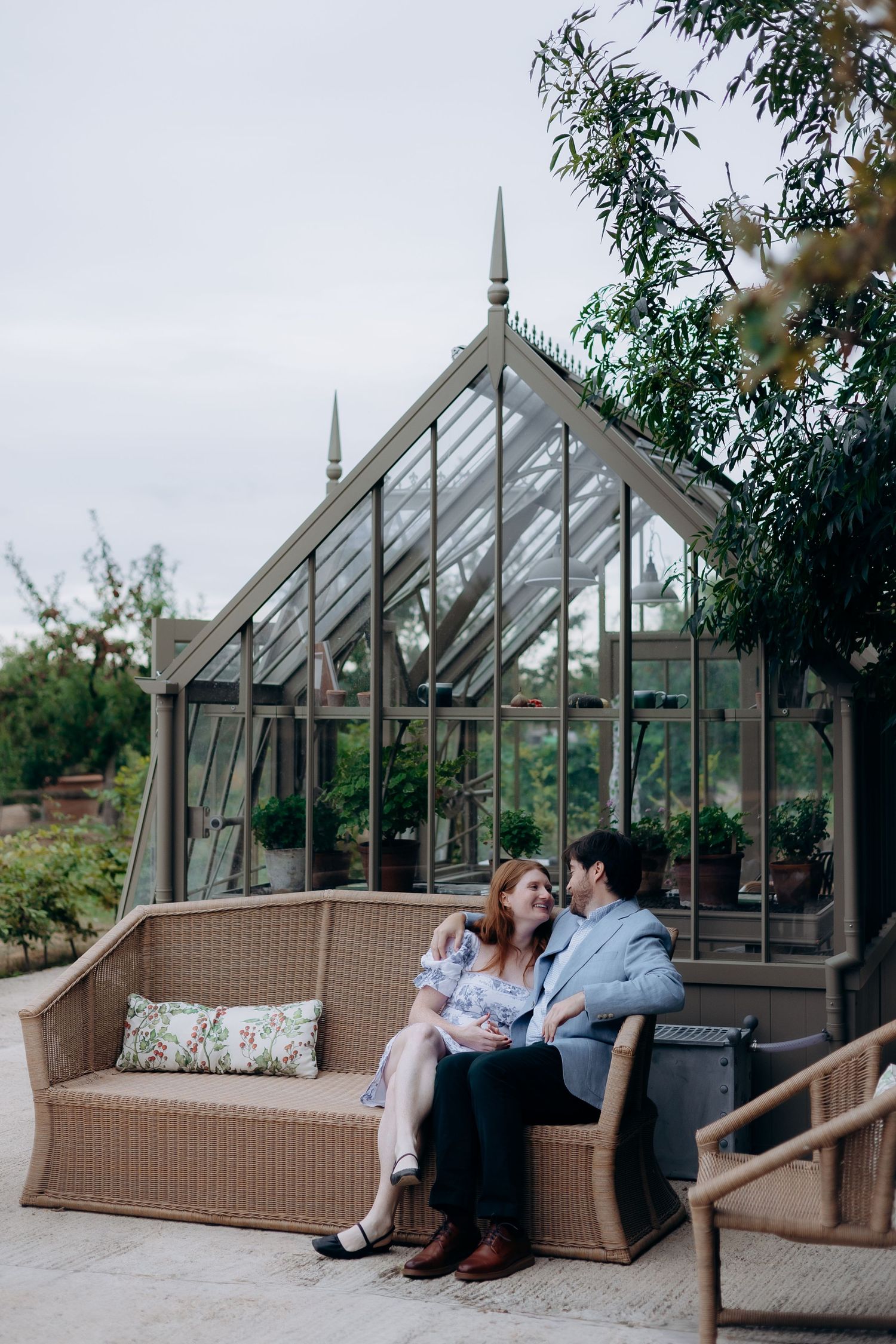 Couple sitting together on wicker sofa in front of elegant glass greenhouse in garden setting.