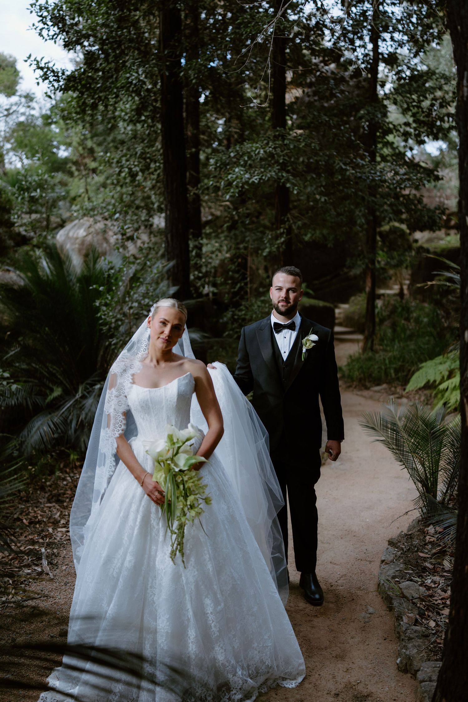 Bride and groom walk through mountain meadow at golden hour, she in white ballgown, he in black tuxedo.