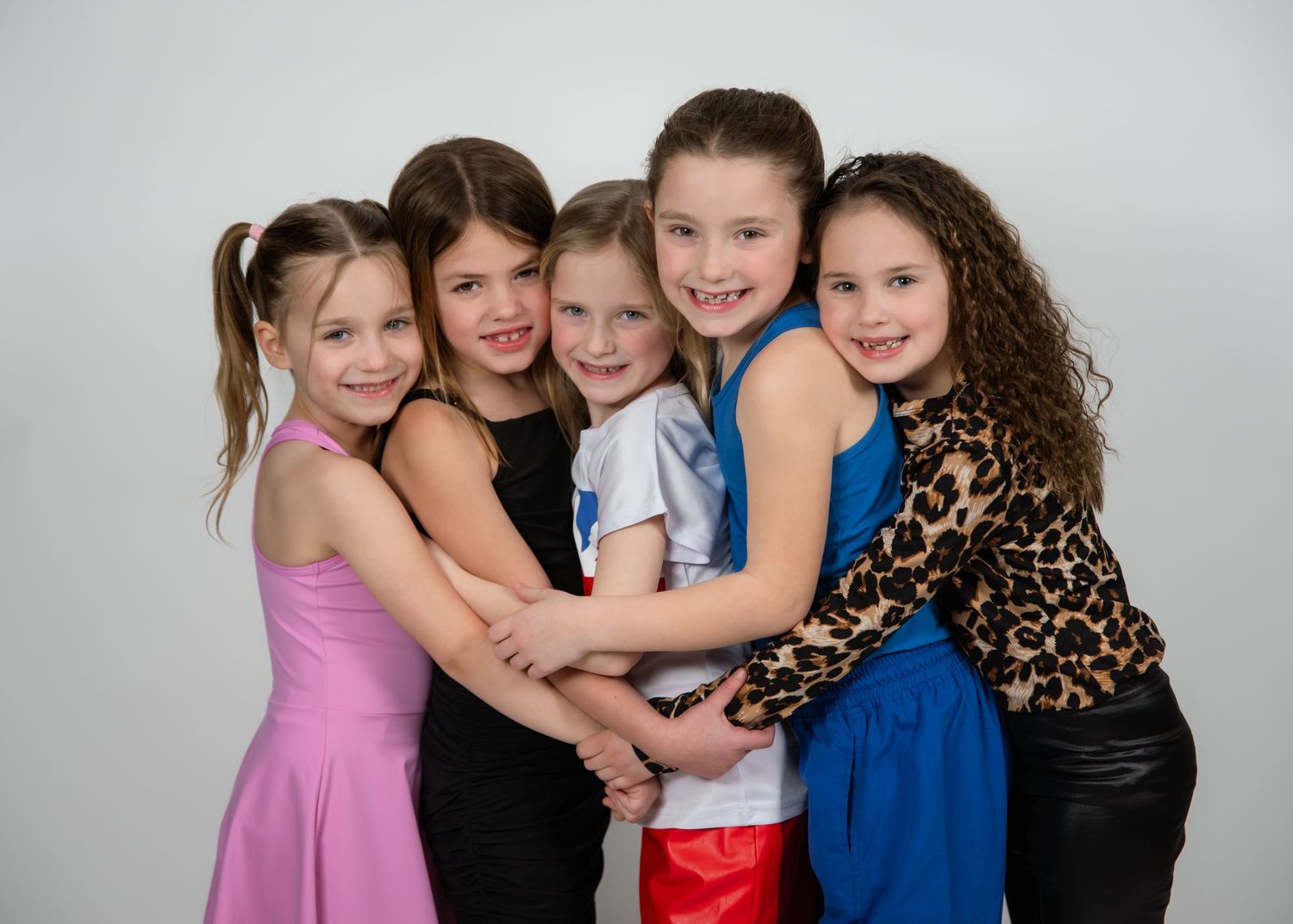 Five young girls in colorful dresses hugging and smiling together in a professional photo studio setting.