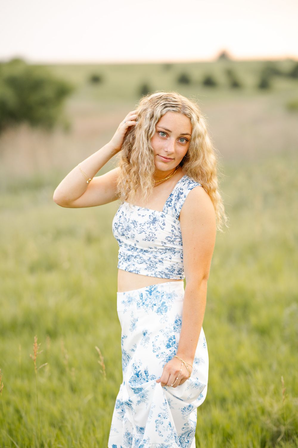 Young blonde woman in blue floral crop top and skirt posing in green meadow at golden hour.