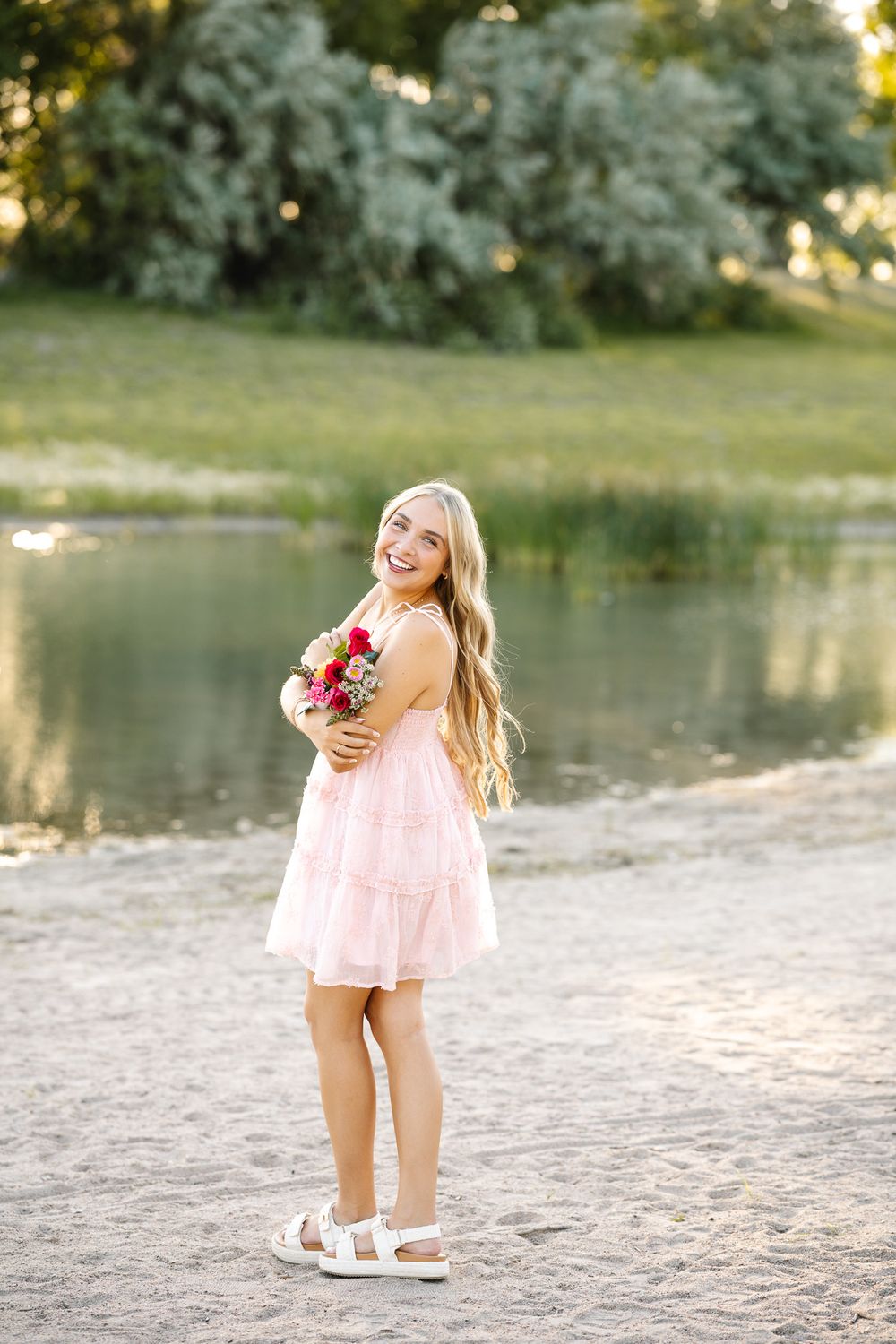 Smiling blonde woman in pink dress holding colorful bouquet by a sunlit lake in a park.