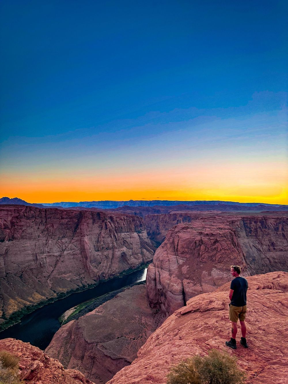 Solo hiker standing on red rock cliff overlooking Horseshoe Bend canyon at stunning sunset.
