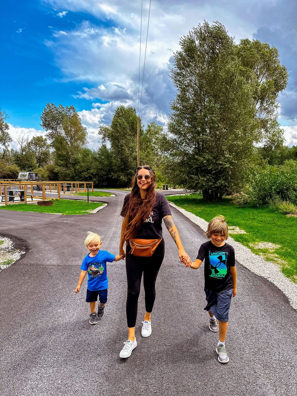 Woman with long hair holding hands with two boys in blue shirts walking on sunny path.