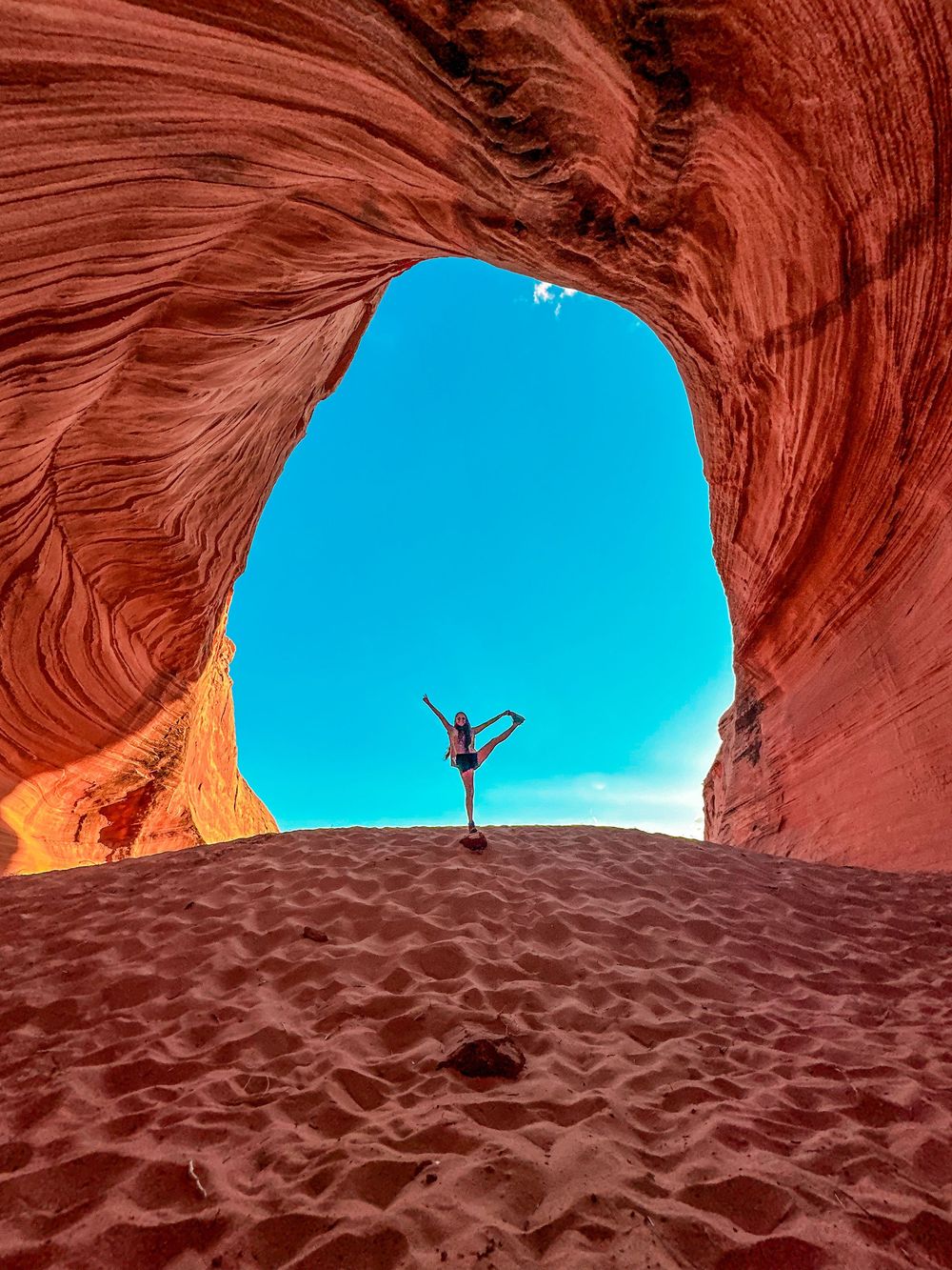 Person striking yoga pose inside massive red sandstone arch cave with blue sky backdrop.