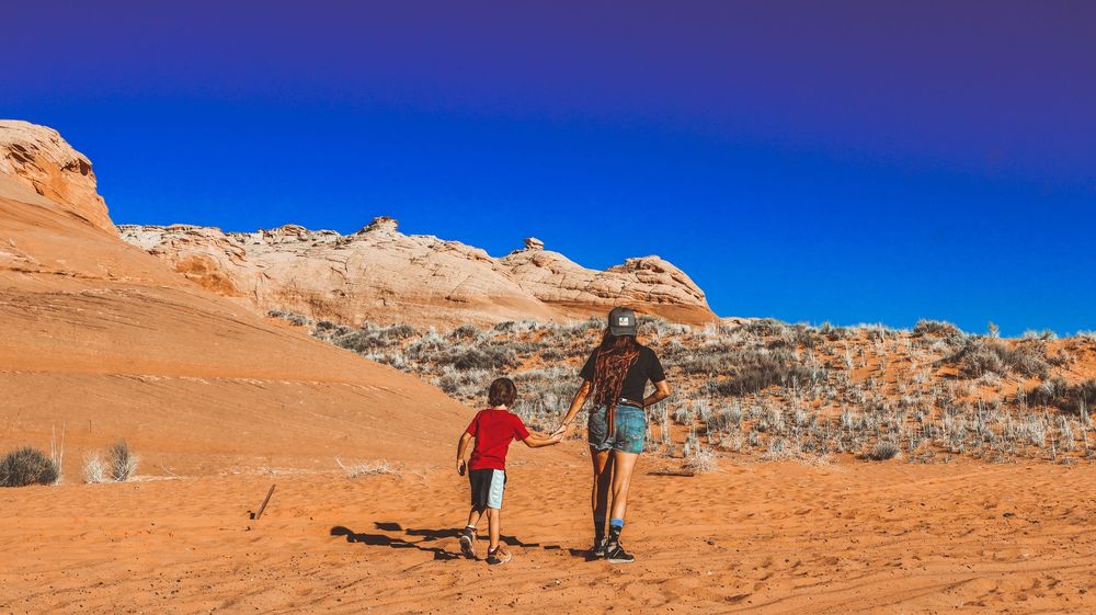 Woman and child walking hand-in-hand toward white rock formations on red sand desert trail.