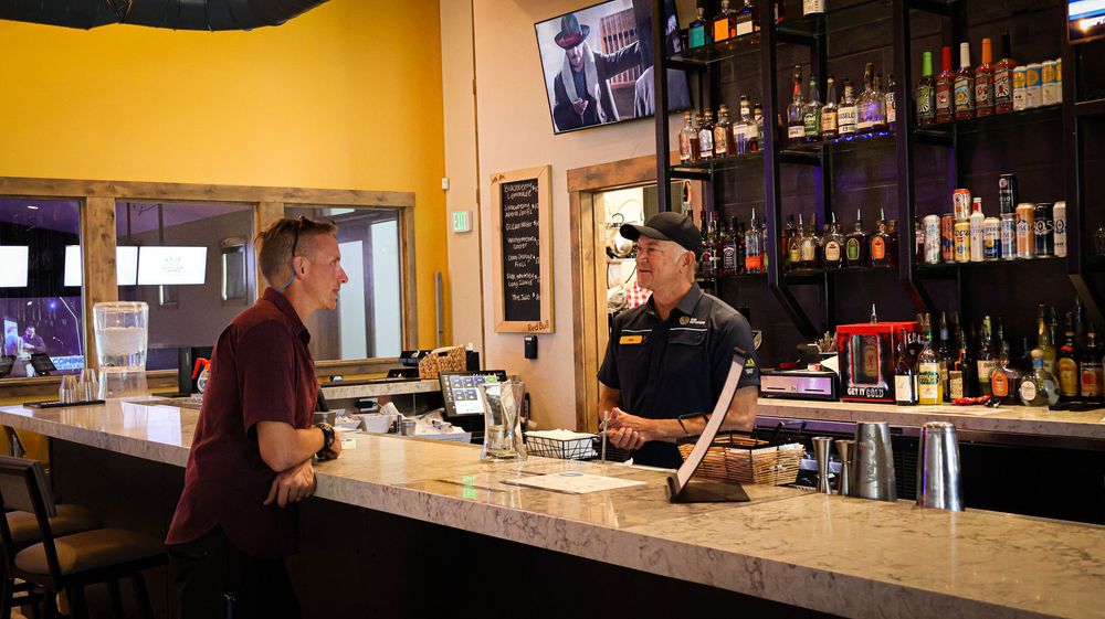 Two men chatting at a bar counter with bottles and TV screens in background.