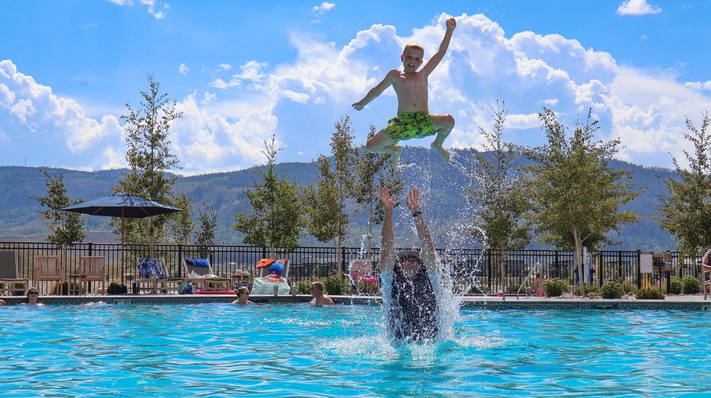 Excited boy jumping high above a swimming pool with mountain views and blue sky backdrop.