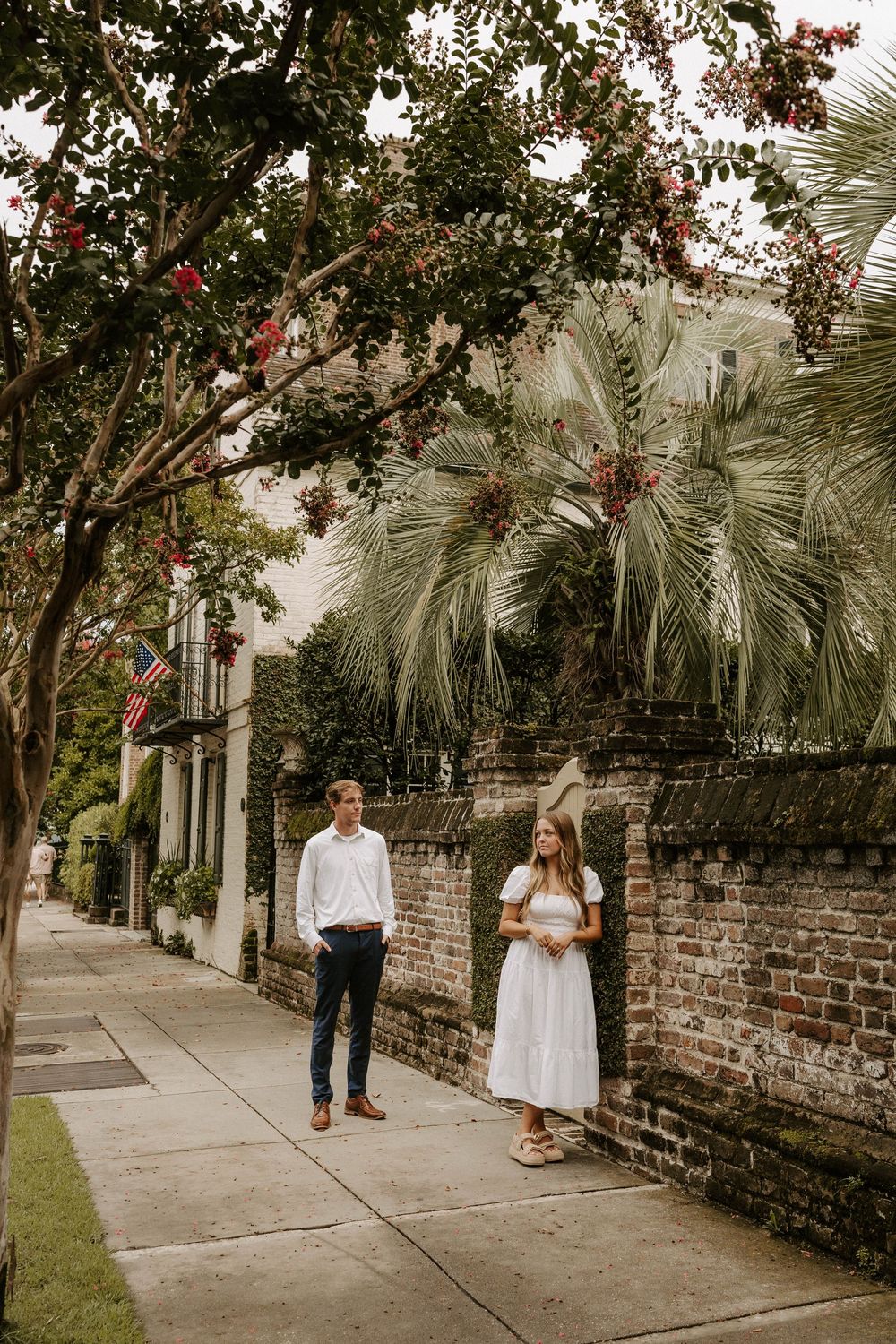 Couple stands apart on a historic Charleston sidewalk with brick wall, palm trees, and American flags in background.